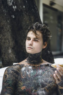 Genç, yakışıklı, dövmeli bir adam kahvaltısını yapıyor açık havada bahçede, açık havada, kahvaltıda çörek ve kahve içiyor..