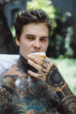 Genç, yakışıklı, dövmeli bir adam kahvaltısını yapıyor açık havada bahçede, açık havada, kahvaltıda çörek ve kahve içiyor..