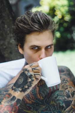 Genç, yakışıklı, dövmeli bir adam kahvaltısını yapıyor açık havada bahçede, açık havada, kahvaltıda çörek ve kahve içiyor..