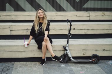 Uzun saçlı, elektrikli scooterlı genç bir kadın. Elektrikli scooterlı kız kahve içiyor..