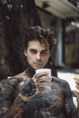 Genç, yakışıklı, dövmeli bir adam kahvaltısını yapıyor açık havada bahçede, açık havada, kahvaltıda çörek ve kahve içiyor..