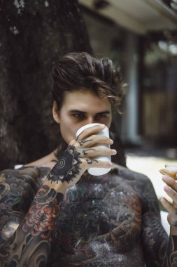 Genç, yakışıklı, dövmeli bir adam kahvaltısını yapıyor açık havada bahçede, açık havada, kahvaltıda çörek ve kahve içiyor..