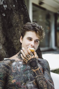 Genç, yakışıklı, dövmeli bir adam kahvaltısını yapıyor açık havada bahçede, açık havada, kahvaltıda çörek ve kahve içiyor..