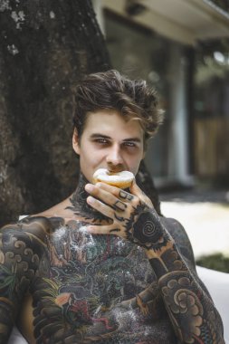 Genç, yakışıklı, dövmeli bir adam kahvaltısını yapıyor açık havada bahçede, açık havada, kahvaltıda çörek ve kahve içiyor..