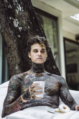 Genç, yakışıklı, dövmeli bir adam kahvaltısını yapıyor açık havada bahçede, açık havada, kahvaltıda çörek ve kahve içiyor..