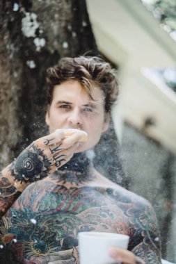Genç, yakışıklı, dövmeli bir adam kahvaltısını yapıyor açık havada bahçede, açık havada, kahvaltıda çörek ve kahve içiyor..