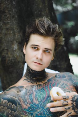 Genç, yakışıklı, dövmeli bir adam kahvaltısını yapıyor açık havada bahçede, açık havada, kahvaltıda çörek ve kahve içiyor..