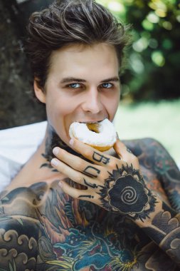 Genç, yakışıklı, dövmeli bir adam kahvaltısını yapıyor açık havada bahçede, açık havada, kahvaltıda çörek ve kahve içiyor..