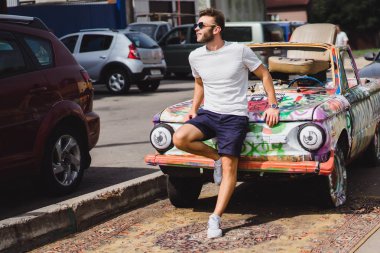 Genç adam eski bir arabanın arka planında poz veriyor.