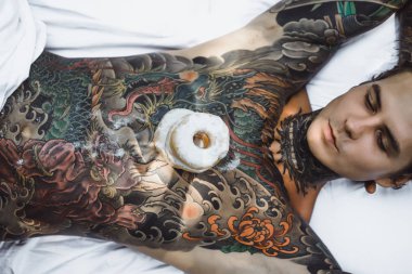 Genç, yakışıklı, dövmeli bir adam kahvaltısını yapıyor açık havada bahçede, açık havada, kahvaltıda çörek ve kahve içiyor..