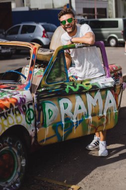 Genç adam eski bir arabanın arka planında poz veriyor.