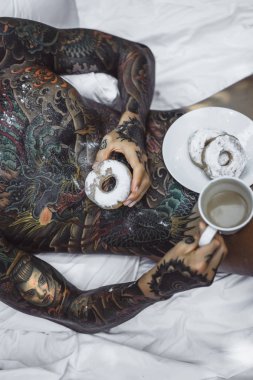 Genç, yakışıklı, dövmeli bir adam kahvaltısını yapıyor açık havada bahçede, açık havada, kahvaltıda çörek ve kahve içiyor..