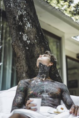 Genç, yakışıklı, dövmeli bir adam kahvaltısını yapıyor açık havada bahçede, açık havada, kahvaltıda çörek ve kahve içiyor..