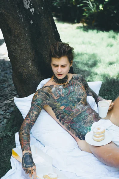 young handsome tattooed man having breakfast in bed in the open air outdoors in the garden, donuts and coffee for breakfast.