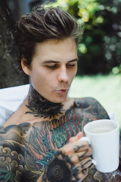 Genç, yakışıklı, dövmeli bir adam kahvaltısını yapıyor açık havada bahçede, açık havada, kahvaltıda çörek ve kahve içiyor..