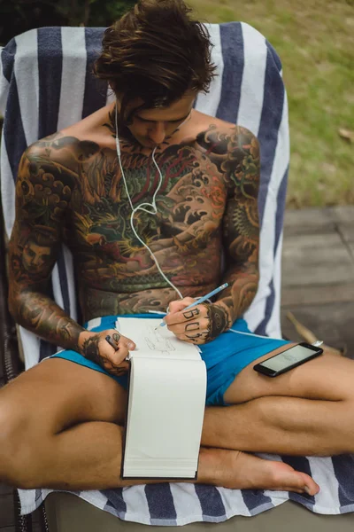 dövmeler kulaklıklar giyen genç bir erkekte müziği dinliyor ve içinde a defter bir kalem çizer. Çevrimiçi eğitim.