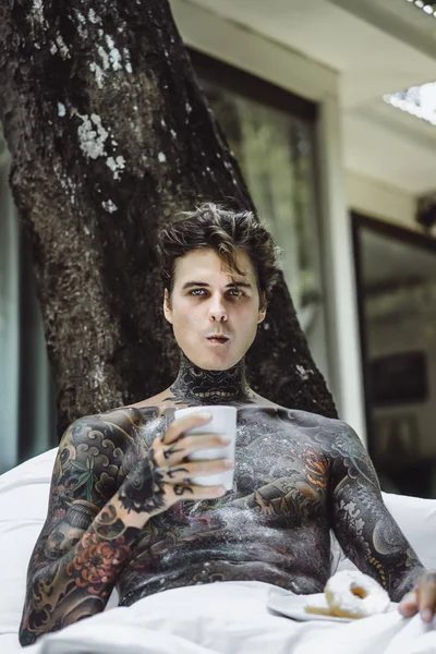 Genç, yakışıklı, dövmeli bir adam kahvaltısını yapıyor açık havada bahçede, açık havada, kahvaltıda çörek ve kahve içiyor..