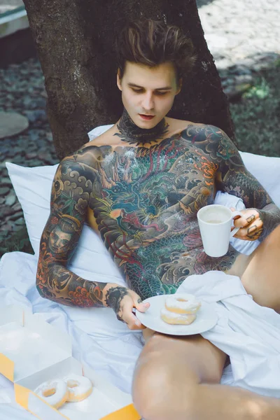 若いハンサムな入れ墨の男は庭の屋外の屋外でベッドで朝食を持っています 朝食のためのドーナツとコーヒー — ストック写真