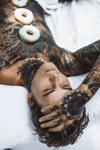 young handsome tattooed man having breakfast in bed in the open air outdoors in the garden, donuts and coffee for breakfast.