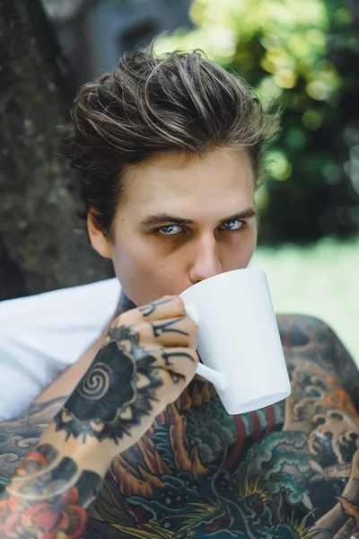 Genç, yakışıklı, dövmeli bir adam kahvaltısını yapıyor açık havada bahçede, açık havada, kahvaltıda çörek ve kahve içiyor..