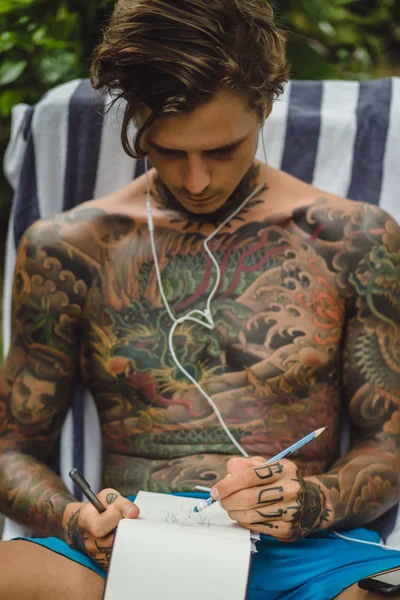 Giovane Tatuato Con Cuffie Ascolta Musica Disegna Una Matita Quaderno — Foto Stock
