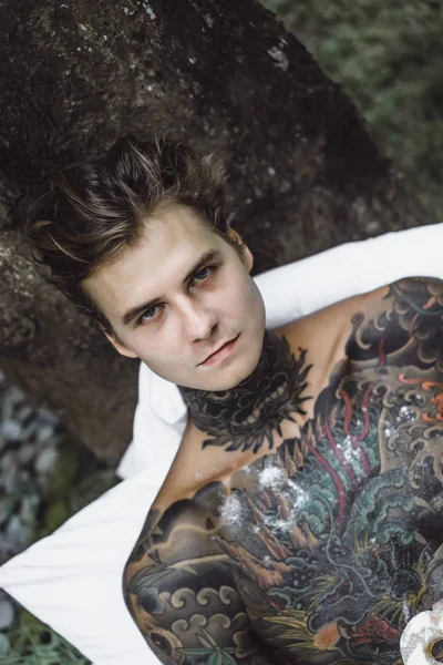 young handsome tattooed man having breakfast in bed in the open air outdoors in the garden, donuts and coffee for breakfast.