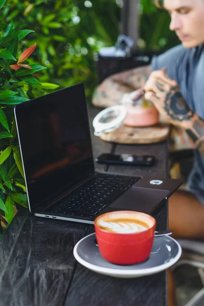 A man in tattoos breakfasts in an outdoor cafe, works on a laptop, drinks coffee. The concept of freelancing