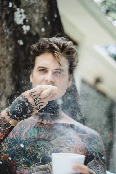 Genç, yakışıklı, dövmeli bir adam kahvaltısını yapıyor açık havada bahçede, açık havada, kahvaltıda çörek ve kahve içiyor..