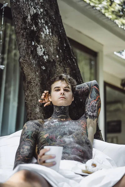 Genç, yakışıklı, dövmeli bir adam kahvaltısını yapıyor açık havada bahçede, açık havada, kahvaltıda çörek ve kahve içiyor..