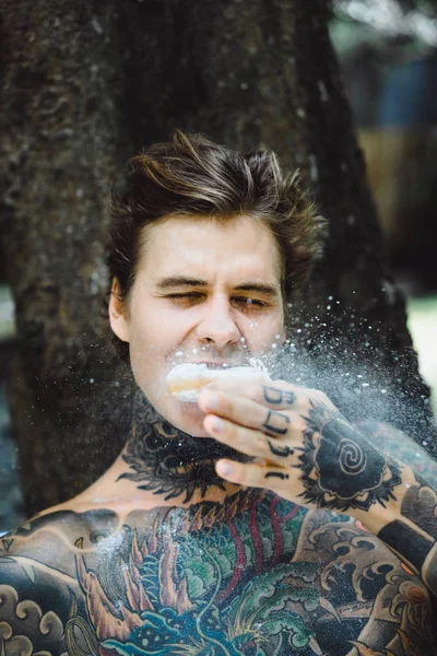 Genç, yakışıklı, dövmeli bir adam kahvaltısını yapıyor açık havada bahçede, açık havada, kahvaltıda çörek ve kahve içiyor..