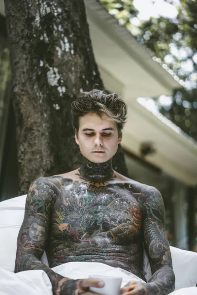 Genç, yakışıklı, dövmeli bir adam kahvaltısını yapıyor açık havada bahçede, açık havada, kahvaltıda çörek ve kahve içiyor..