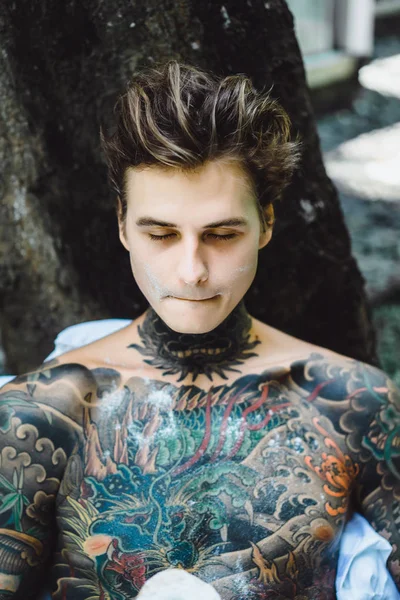 Genç, yakışıklı, dövmeli bir adam kahvaltısını yapıyor açık havada bahçede, açık havada, kahvaltıda çörek ve kahve içiyor..