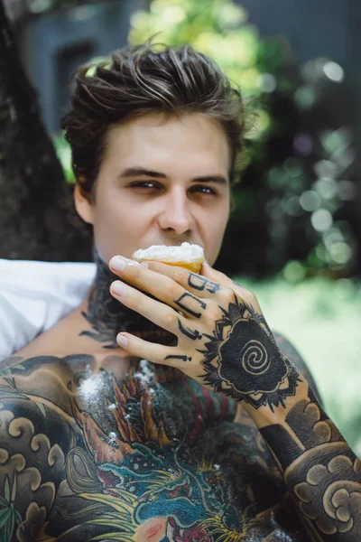 Genç, yakışıklı, dövmeli bir adam kahvaltısını yapıyor açık havada bahçede, açık havada, kahvaltıda çörek ve kahve içiyor..