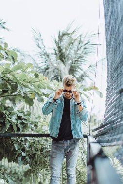 tropikal bir yerde, palmiye ağaçları ve yeşil, güneş gözlüklü bir arka plan üzerinde poz genç çekici adam