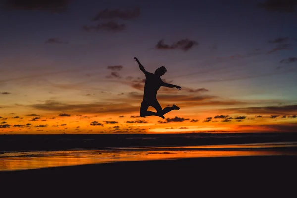 夕暮れ時の海の海岸に人々 男が夕日を背にジャンプします — ストック写真