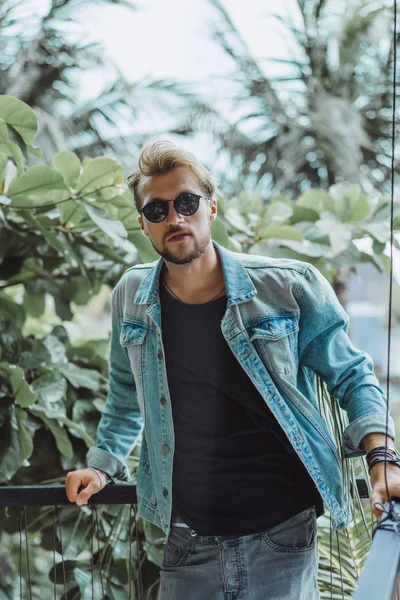 tropikal bir yerde, palmiye ağaçları ve yeşil, güneş gözlüklü bir arka plan üzerinde poz genç çekici adam