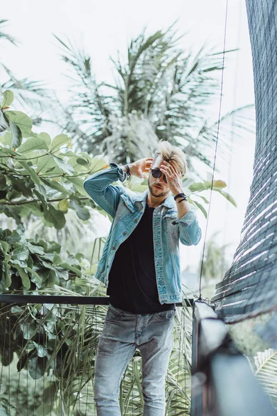 tropikal bir yerde, palmiye ağaçları ve yeşil, güneş gözlüklü bir arka plan üzerinde poz genç çekici adam