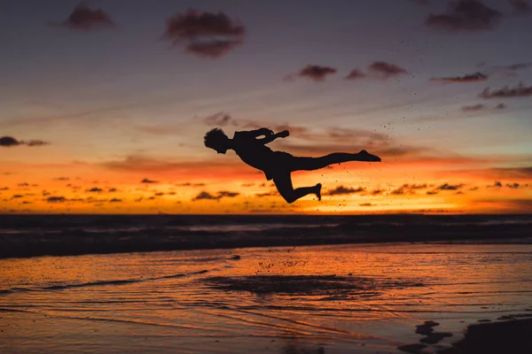 夕暮れ時の海の海岸に人々 男が夕日を背にジャンプします — ストック写真