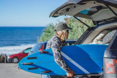 bir kap ve bir surfboard ile güneş gözlüğü uzun beyaz saçlı sörfçü adam. sörf sonra döndürür, arabaya sörf tahtası kıvrımları. Nazare, Portekiz.