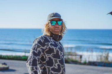 bir kap ve bir surfboard ile güneş gözlüğü uzun beyaz saçlı sörfçü adam. sörf sonra döndürür, arabaya sörf tahtası kıvrımları. Nazare, Portekiz.