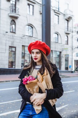 Genç güzel kız Fransız kadın esmer kırmızı bere ve siyah bir ceket Avrupa şehri Street'te bir buket çiçek ve Fransız baget ile gider. Mutlu bir gülümseme ve büyüleyici bir bak, Paris Fransız tarzı
