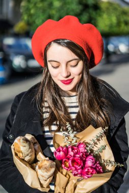 Genç güzel kız Fransız kadın esmer kırmızı bere ve siyah bir ceket Avrupa şehri Street'te bir buket çiçek ve Fransız baget ile gider. Mutlu bir gülümseme ve büyüleyici bir bak, Paris Fransız tarzı