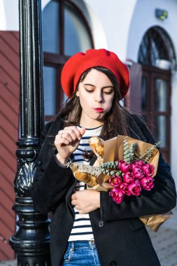 Genç güzel kız Fransız kadın esmer kırmızı bere ve siyah bir ceket Avrupa şehri Street'te bir buket çiçek ve Fransız baget ile gider. Mutlu bir gülümseme ve büyüleyici bir bak, Paris Fransız tarzı