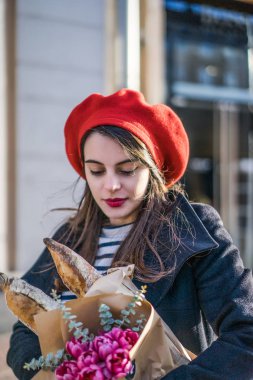 Genç güzel kız Fransız kadın esmer kırmızı bere ve siyah bir ceket Avrupa şehri Street'te bir buket çiçek ve Fransız baget ile gider. Mutlu bir gülümseme ve büyüleyici bir bak, Paris Fransız tarzı