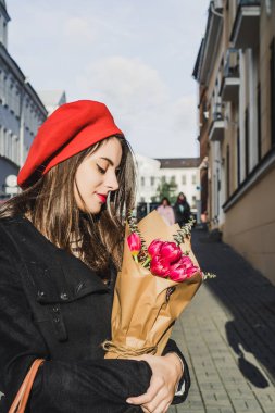 Genç güzel kız Fransız kadın esmer kırmızı bere ve siyah bir ceket Avrupa şehri Street'te bir buket çiçek ve Fransız baget ile gider. Mutlu bir gülümseme ve büyüleyici bir bak, Paris Fransız tarzı
