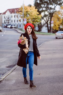 Genç güzel kız Fransız kadın esmer kırmızı bere ve siyah bir ceket Avrupa şehri Street'te bir buket çiçek ve Fransız baget ile gider. Mutlu bir gülümseme ve büyüleyici bir bak, Paris Fransız tarzı