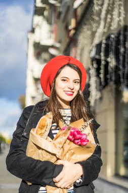 Genç güzel kız Fransız kadın esmer kırmızı bere ve siyah bir ceket Avrupa şehri Street'te bir buket çiçek ve Fransız baget ile gider. Mutlu bir gülümseme ve büyüleyici bir bak, Paris Fransız tarzı