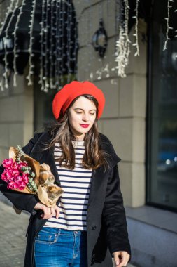 Genç güzel kız Fransız kadın esmer kırmızı bere ve siyah bir ceket Avrupa şehri Street'te bir buket çiçek ve Fransız baget ile gider. Mutlu bir gülümseme ve büyüleyici bir bak, Paris Fransız tarzı