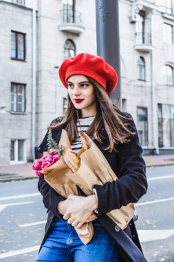 Genç güzel kız Fransız kadın esmer kırmızı bere ve siyah bir ceket Avrupa şehri Street'te bir buket çiçek ve Fransız baget ile gider. Mutlu bir gülümseme ve büyüleyici bir bak, Paris Fransız tarzı