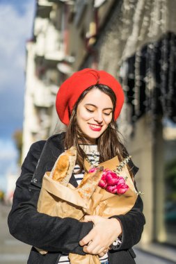 Genç güzel kız Fransız kadın esmer kırmızı bere ve siyah bir ceket Avrupa şehri Street'te bir buket çiçek ve Fransız baget ile gider. Mutlu bir gülümseme ve büyüleyici bir bak, Paris Fransız tarzı